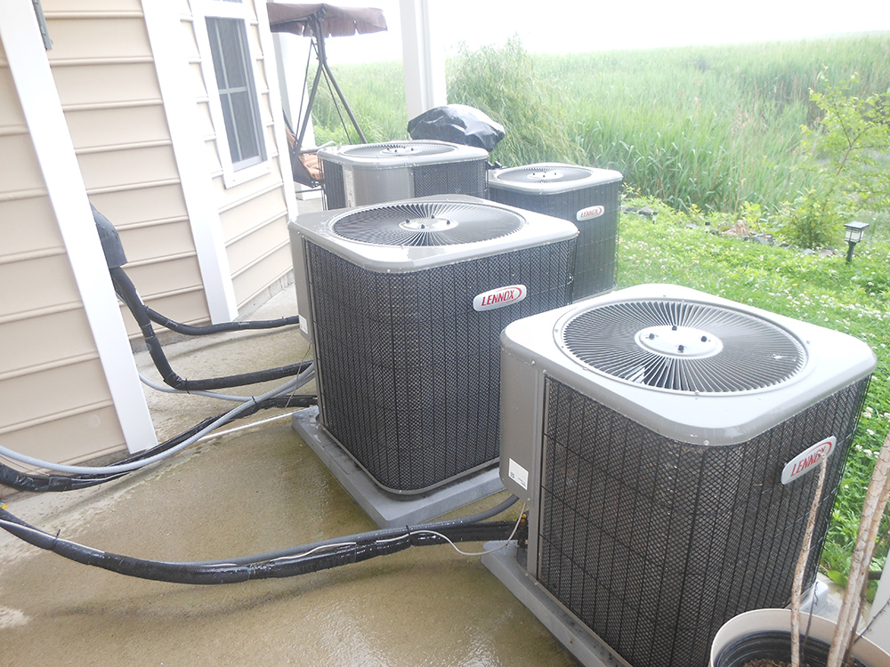 Four outdoor central air conditioning units on a concrete patio next to a house, surrounded by greenery.