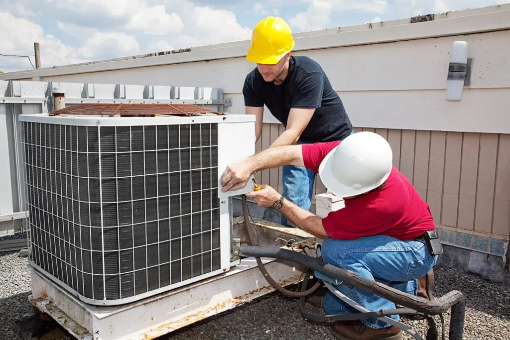 Technicians repairing rooftop HVAC unit, wearing safety helmets, performing air conditioning maintenance under clear sky.