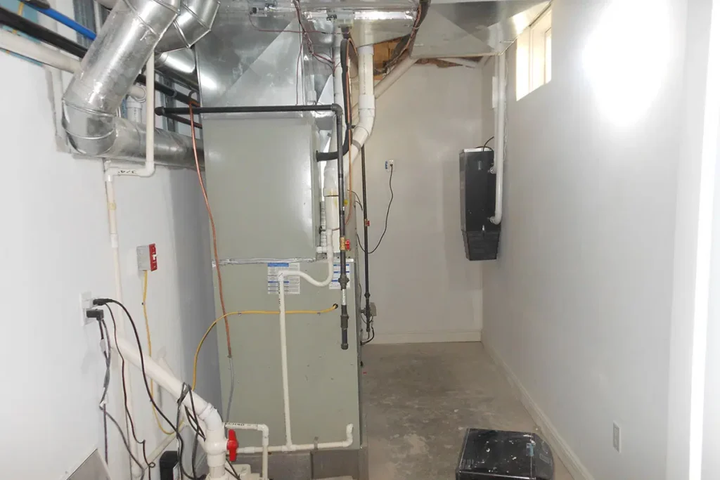 Basement furnace room with HVAC system, ductwork, and pipes in a narrow hallway with a small window.