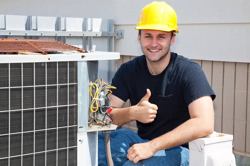 Smiling HVAC technician in yellow hard hat gives thumbs up next to air conditioning unit, showcasing repair service.