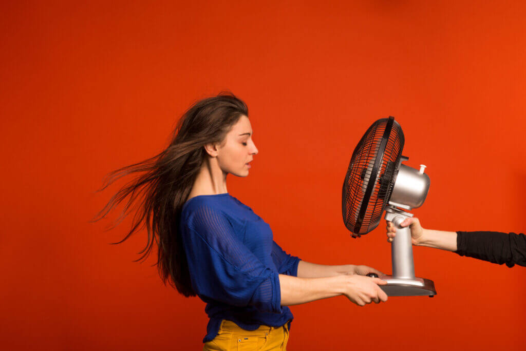 Young woman holding a fan, hair blowing, against vibrant orange backdrop in studio portrait.