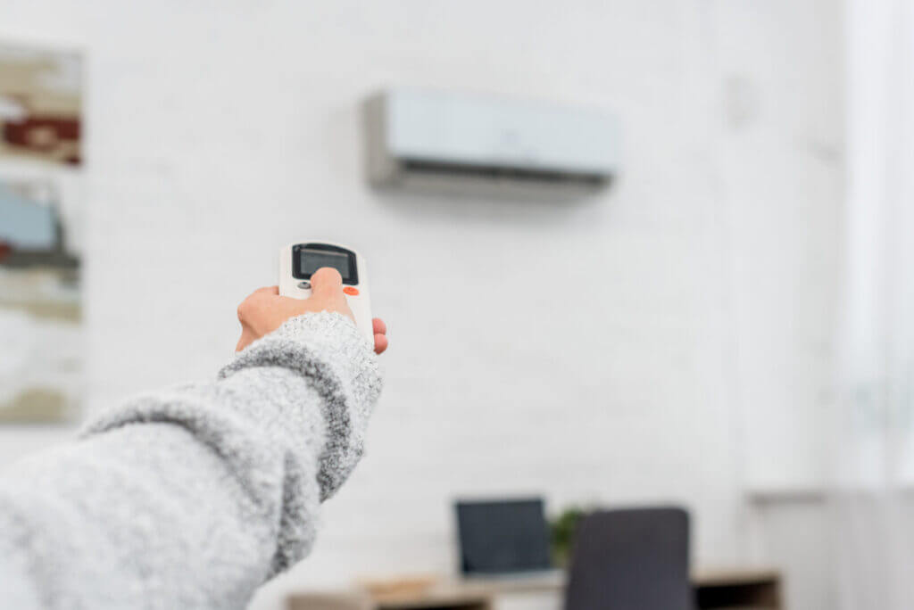 Woman in sweater using remote control to adjust wall-mounted air conditioner in modern home office.