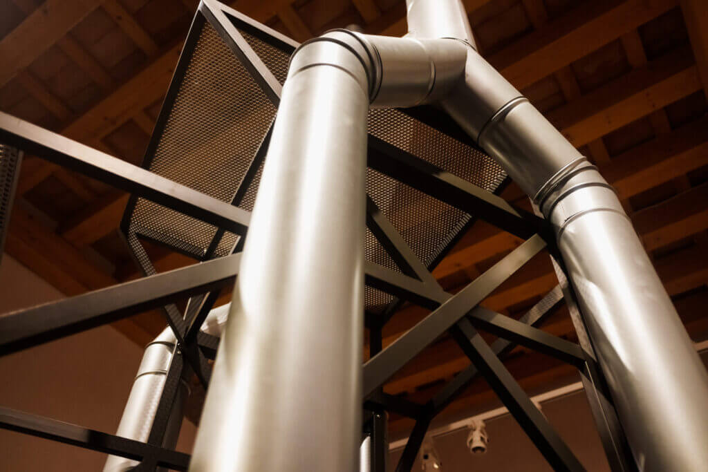 Vertical metal pipes in industrial warehouse, showcasing structural design and metallic framework against wooden ceiling.